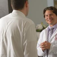 A photograph of a Fox Chase doctor smiling widely at someone, with another person on the phone at a desk to their left.