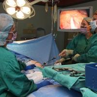 A photograph of three doctors performing surgery, looking up at two screens showing the feed from a laparoscope.