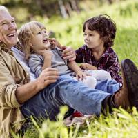 A photograph of an older man sitting in a grassy field with two children on his lap, all smiling widely.