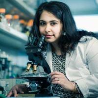 A photograph of Dr. Sanjeevani Arora, PhD, holding a microscope close to her and looking up at the camera.