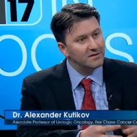 Dr. Alexander Kutikov on a TV show with the channel's logo in the background, speaking to someone off camera.