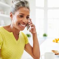 A photo of a person standing in their kitchen with a phone up to their ear and a laptop in front of them.