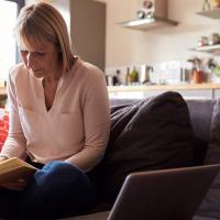 A photo of a woman sitting on the couch with a laptop next to her, holding a pen and pad of paper in her lap.