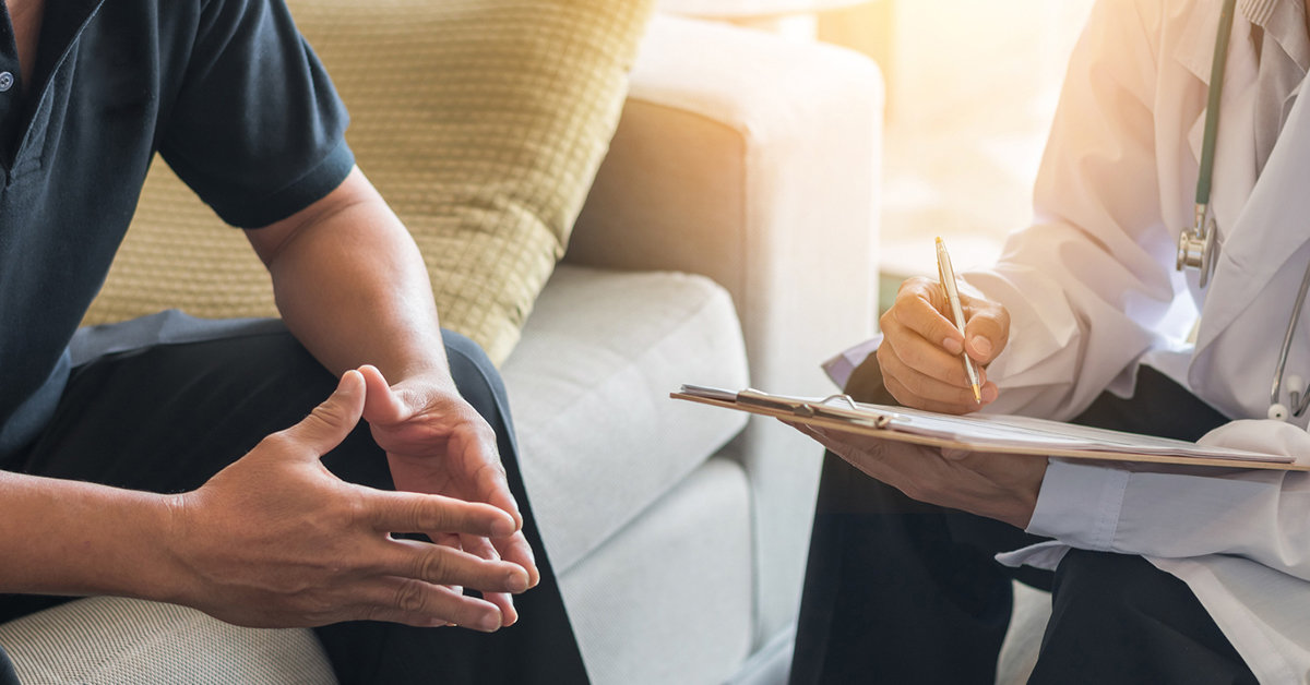 A photograph of a medical professional sitting across from a patient, taking notes on a clipboard.