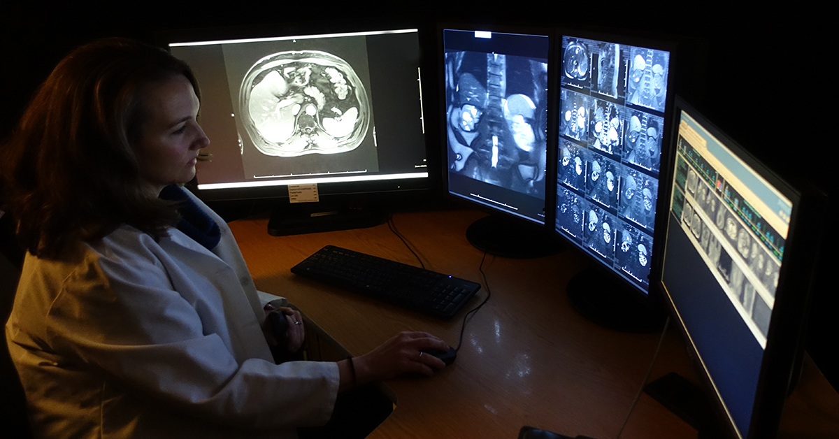 A photograph of a medical professional in a dark room, looking at four screens displaying various medical imaging results.